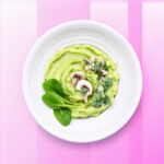 Een smakelijk gerecht van romige avocado-aardappelpuree gegarneerd met verse spinazie en champignons, gepresenteerd op een wit bord tegen een gestreepte roze en witte achtergrond.