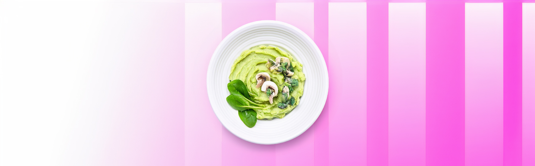 Een smakelijk gerecht van romige avocado-aardappelpuree gegarneerd met verse spinazie en champignons, gepresenteerd op een wit bord tegen een gestreepte roze en witte achtergrond.