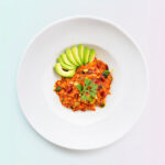 Banner van een Smaakvolle Keto Chili met Avocado en Courgette, smakelijk geserveerd in een witte schaal, geaccentueerd tegen een roze en groene achtergrond.